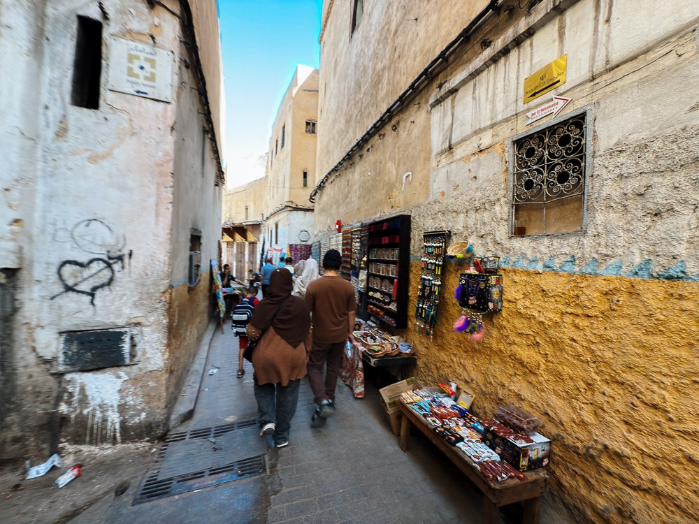 Lorong sempit medina Fez dengan kedai kecil di kiri kanan, laluan pejalan kaki menuju kawasan penyamakan