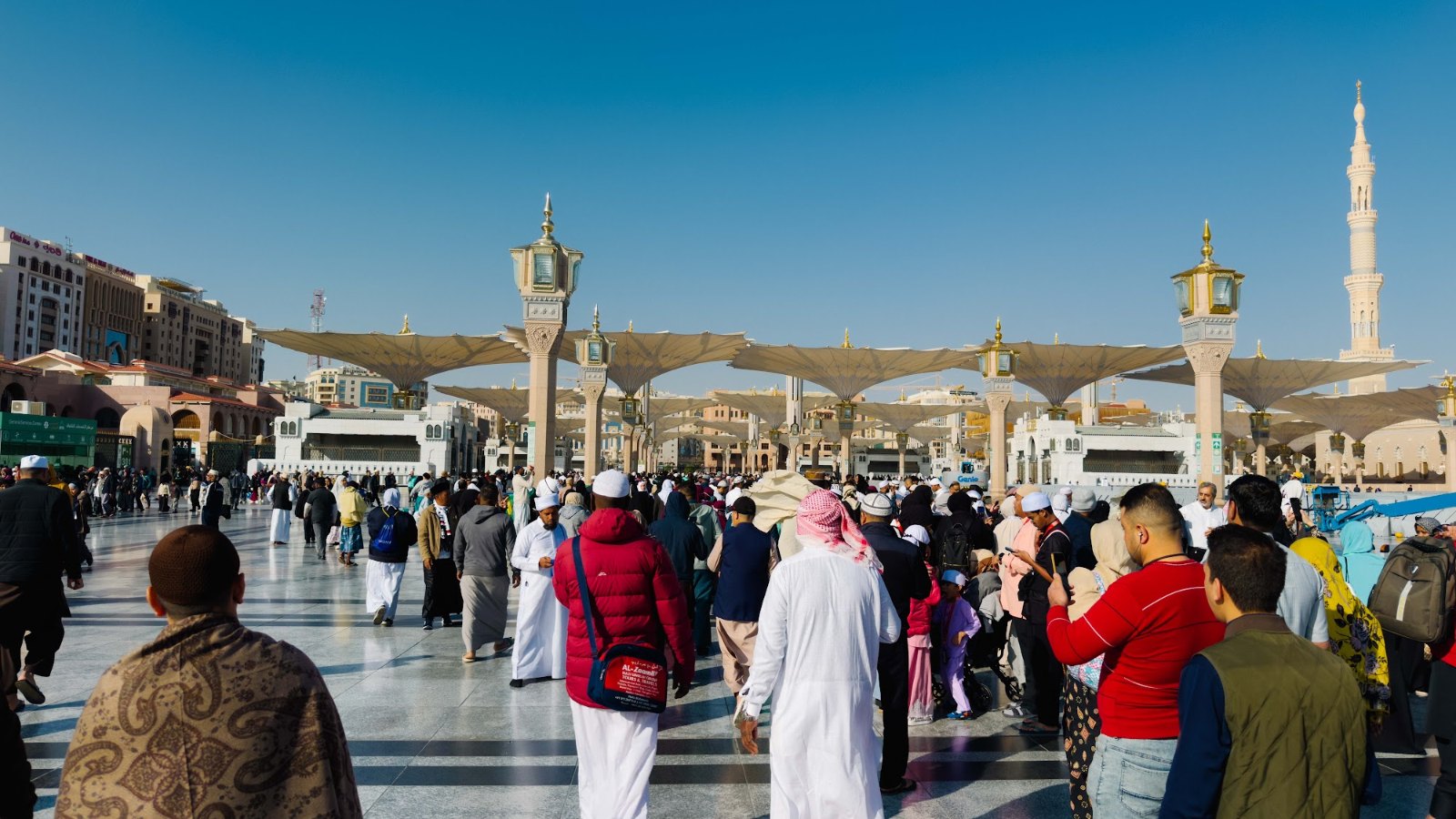 Jemaah pelbagai bangsa berjalan di dataran Masjid Nabawi dengan menara dan payung di latar belakang