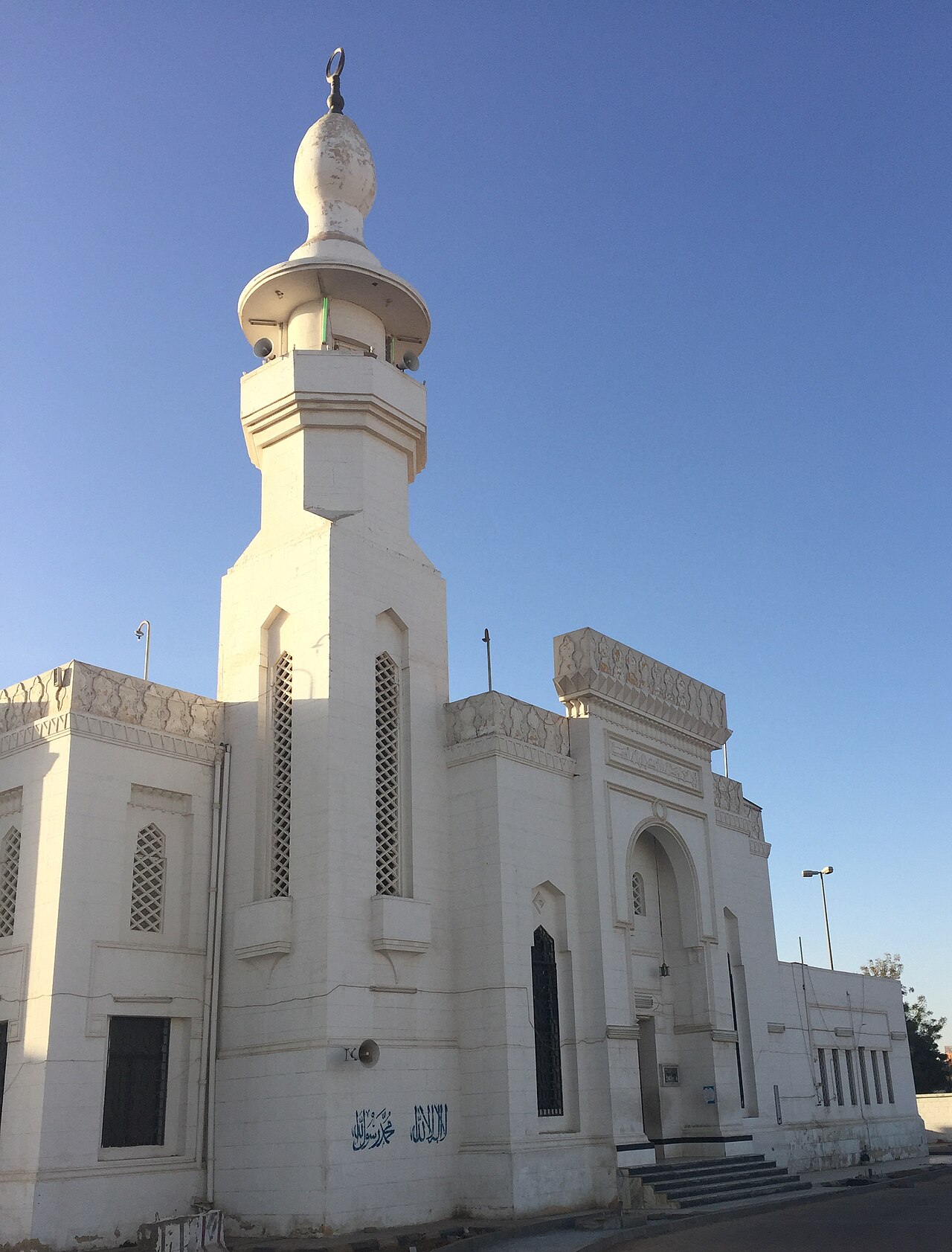 Menara putih Masjid at-Taubah Tabuk dengan dinding berhias geometri di bawah langit biru