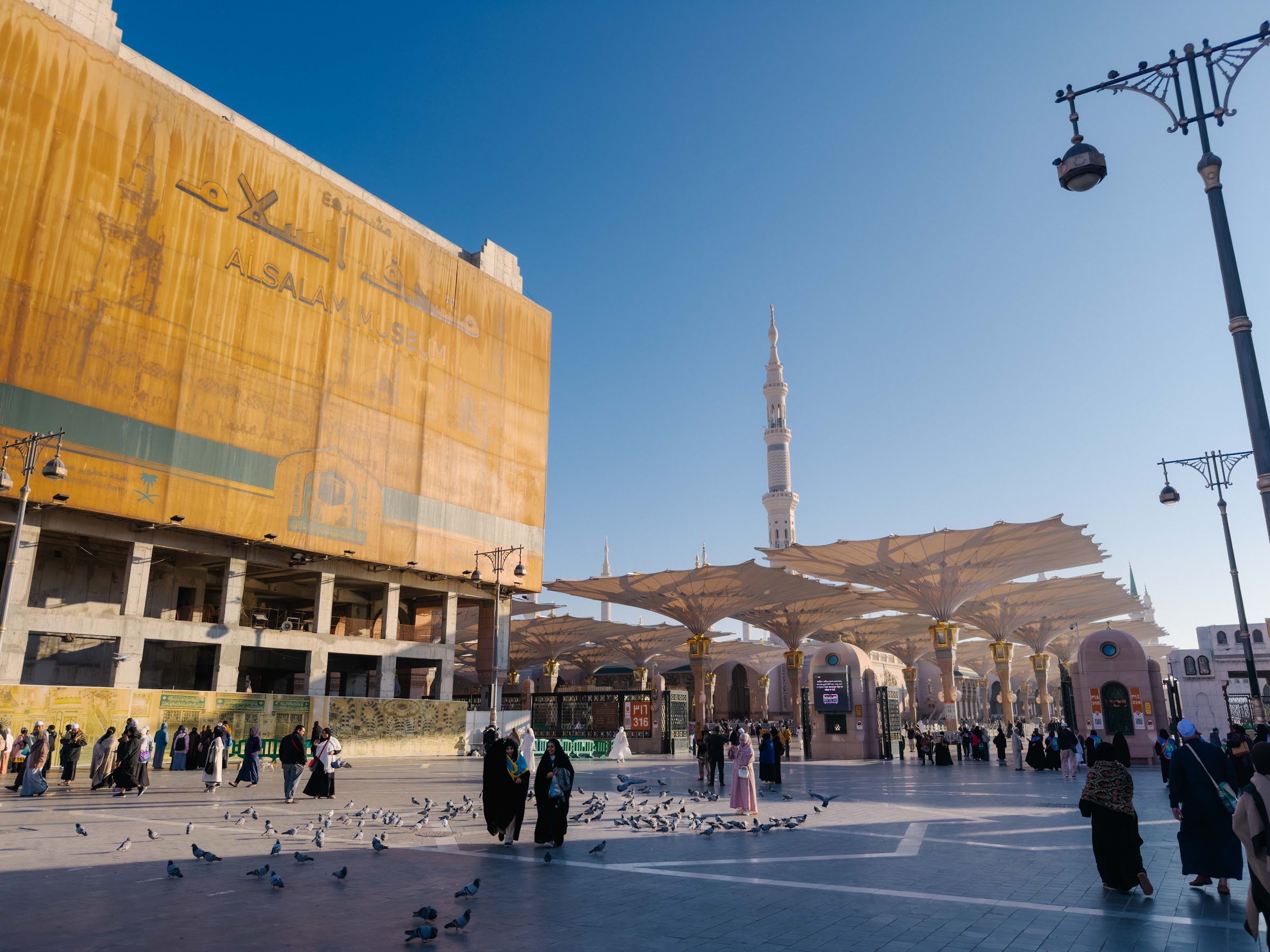Payung gergasi dan menara Masjid Nabawi di halaman masjid pada waktu pagi, dengan jemaah berjalan di dataran