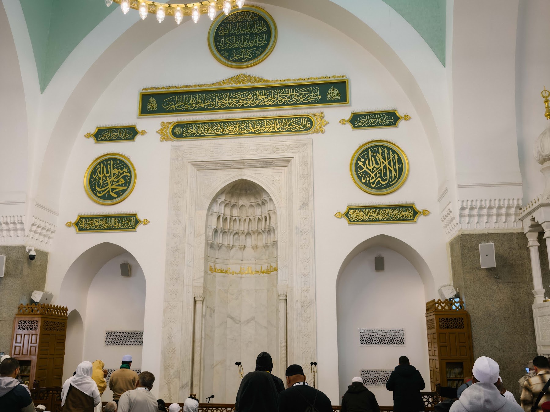 Mihrab Masjid Quba berhias kaligrafi hijau dan emas pada dinding kiblat, dengan jemaah di saf hadapan