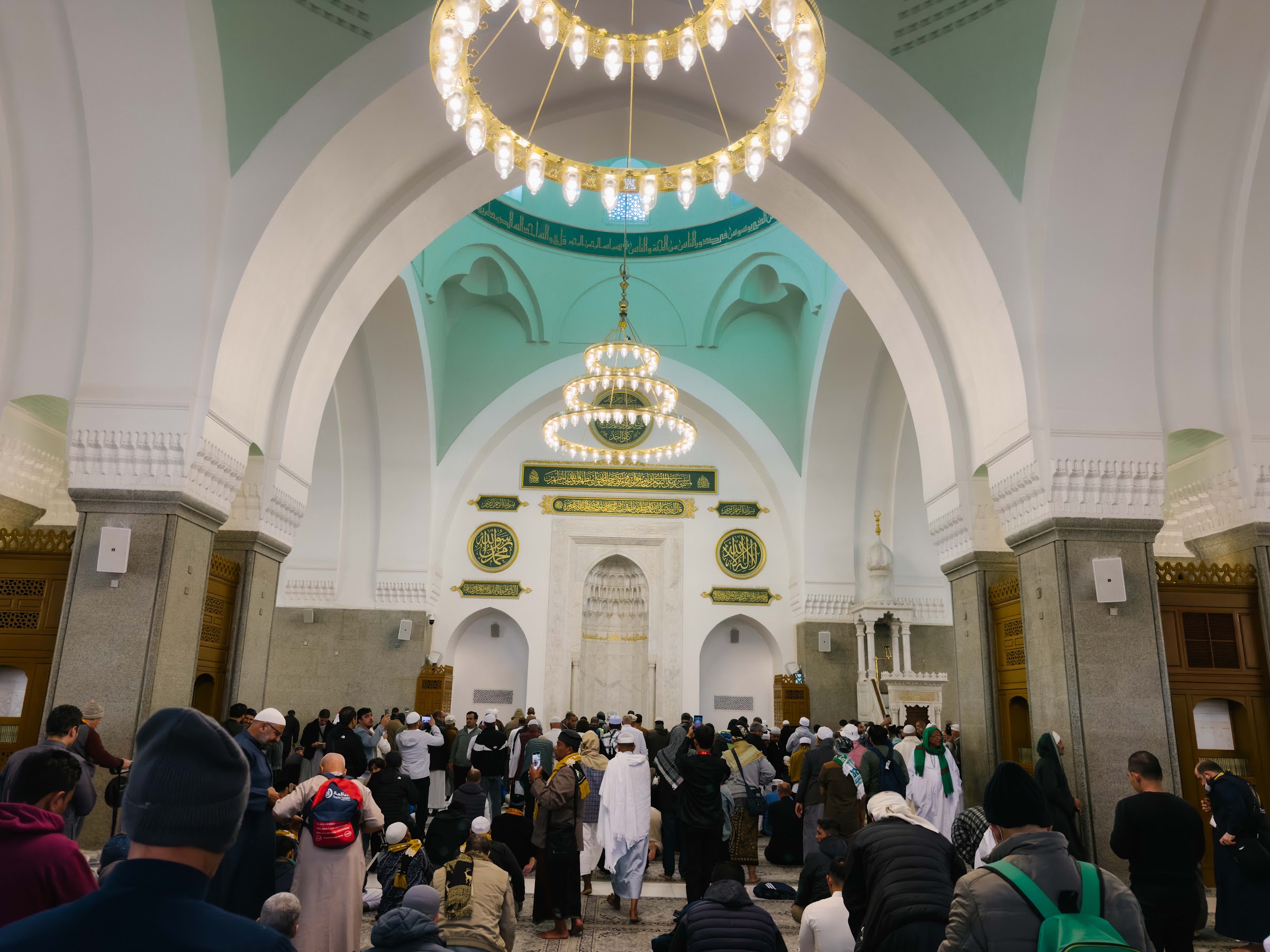 Ruang solat utama Masjid Quba dengan candelier dan kubah hijau muda, dipenuhi jemaah pada waktu pagi