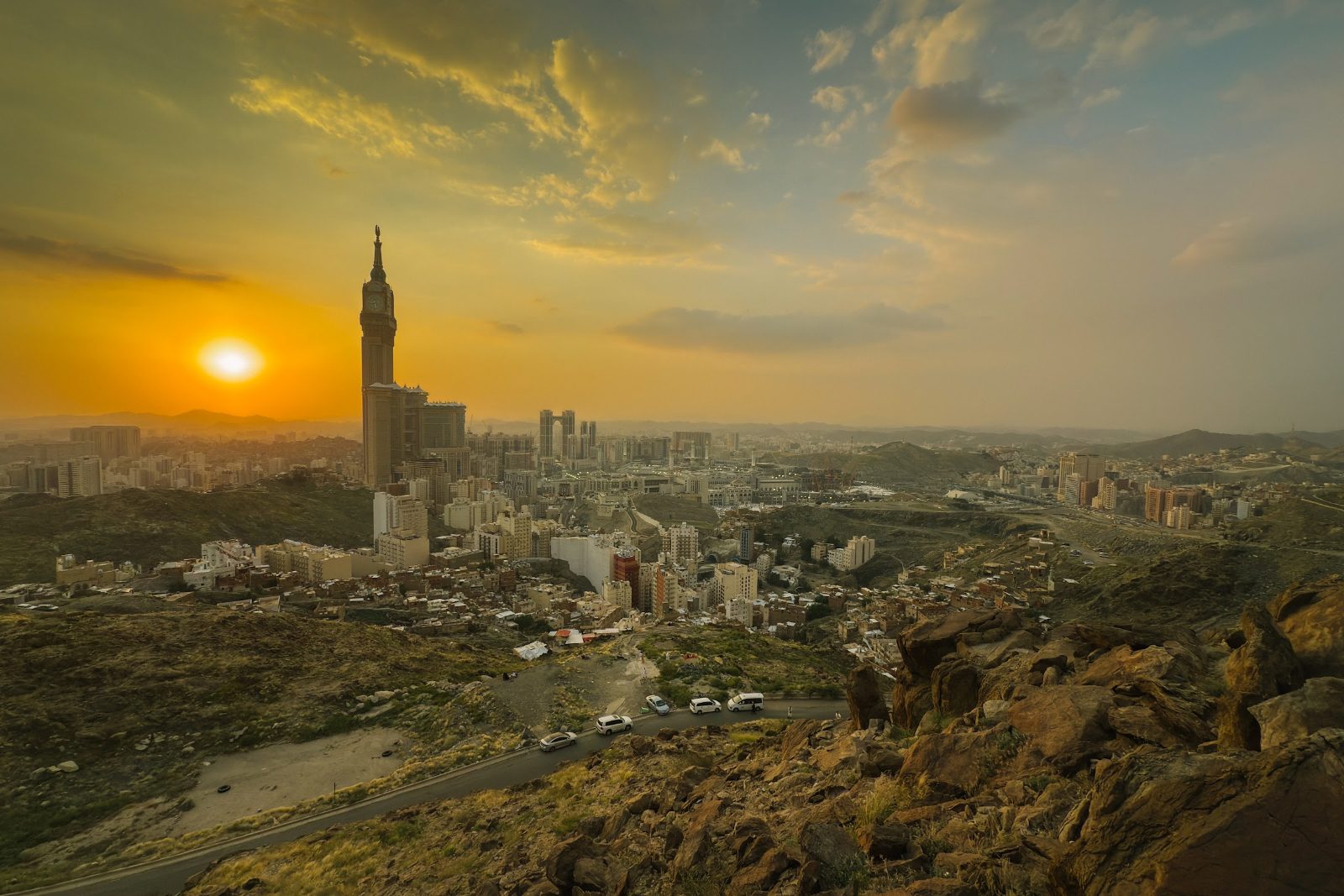 Panorama kota Makkah dari lereng bukit ketika matahari terbenam, dengan menara jam di kejauhan